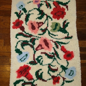 Op de afbeelding: Een wit tapijt met een bloemenpatroon. Het tapijt heeft roze, rode, groene en blauwe bloemen met bruine stelen. Het tapijt is gemaakt van een zacht, pluche materiaal.