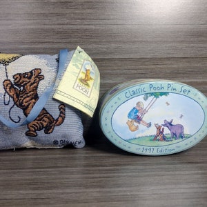 May include: A blue and brown Winnie the Pooh themed cushion with a blue strap and a tag that says "Classic Pooh". Next to the cushion is a round tin with a blue and green Winnie the Pooh design and the text "Classic Pooh Pin Set - 1997 Edition".