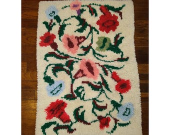 May include: A rectangular, off-white rug with a floral design. The rug features red, pink, blue, and green flowers and leaves. The flowers are densely packed and arranged in a swirling pattern. The rug is made of a soft, textured material.