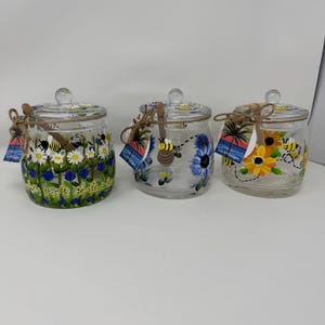 May include: Three clear glass jars with lids, each adorned with hand-painted floral and bee designs. One jar features daisies and blue flowers, another blue flowers, and the third sunflowers. Each jar has a small tag tied with twine.