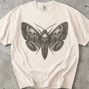 May include: An ivory short-sleeve t-shirt featuring a dark brown graphic print of a moth with a skull on its thorax and two additional skulls integrated into the patterns on its wings. The word IVORY is printed in the upper left corner.