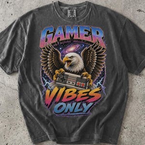 May include: A charcoal gray short-sleeve t-shirt featuring a graphic of a bald eagle holding a retro video game controller against a space background. The text GAMER VIBES ONLY is printed in a colorful gradient font above and below the eagle.
