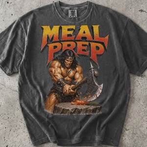 Peut inclure: Un t-shirt gris anthracite à manches courtes avec une impression graphique représentant un guerrier musclé tenant une grande hache de combat au-dessus d'une souche en bois avec une carotte. Le texte MEAL PREP est imprimé en lettres orange et jaune.