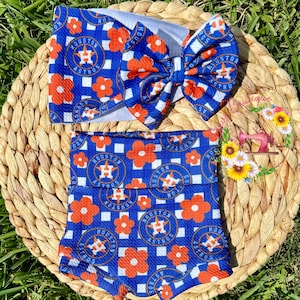 May include: A blue and white headband and shorts set with a floral and Houston Astros logo pattern. The set includes a large bow and shorts, both featuring the team's logo and orange flowers.