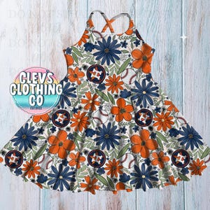 Puede incluir: Vestido sin mangas con un estampado floral de flores naranjas y azul marino, pelotas de béisbol y logotipos de los Astros de Houston. El vestido tiene la espalda cruzada y una falda acampanada. El fondo es blanco.