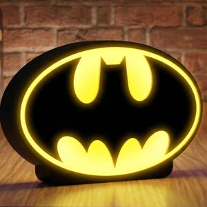 Könnte beinhalten: Eine ovale Batman-Logo-Lampe mit einem schwarzen Rahmen und einer hell leuchtenden gelben Fledermaus-Silhouette in der Mitte. Das Licht strahlt warm aus dem ausgeschnittenen Design heraus und erzeugt einen starken Kontrast zwischen Schwarz und Gelb.