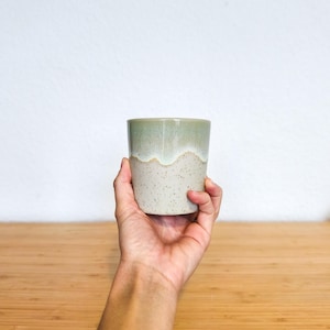 Peut inclure: Une tasse en céramique faite à la main tenue dans une main. La tasse a un design bicolore avec un dessus vert clair et un fond gris clair moucheté. Le bord a un bord ondulé et organique. La tasse est cylindrique et semble non émaillée.