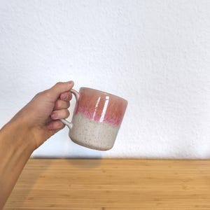 May include: A handmade ceramic mug with a two-tone design. The top half is a warm coral color, transitioning to a speckled beige at the bottom. The mug has a curved handle and is held in a person's hand.