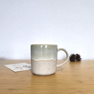 Op de afbeelding: Een keramische mok met een gespikkelde, tweekleurige afwerking. De bovenste helft is een gedempt groen, en de onderste helft is gespikkeld gebroken wit. De mok heeft een afgeronde handgreep en staat op een houten oppervlak. Een kleine kaart en dennenappels zijn ook zichtbaar.