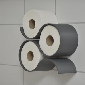Può includere: Un portarotolo da parete grigio scuro con un design curvo a forma di onda che contiene tre rotoli di carta igienica bianca. Il supporto è realizzato in materiale solido e presenta tre aperture circolari per riporre i rotoli di carta.