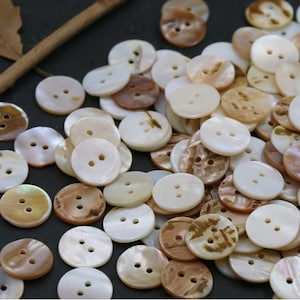 May include: A collection of round, white and brown shell buttons with two holes each. The buttons are arranged in a random pattern on a dark background.