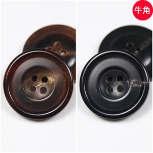 Może przedstawiać: Dwa guziki z czterema otworami, jeden brązowy, a drugi czarny. Guziki są wykonane z rogu i mają gładkie, polerowane wykończenie.