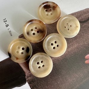 Peut inclure: Six boutons bruns et beiges à quatre trous chacun. Les boutons sont disposés en demi-cercle sur une surface en tissu brun. Le texte "12-B L Size" est visible dans le coin supérieur gauche de l'image.