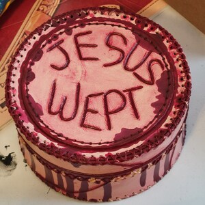 May include: A round, pink leather box with the words "JESUS WEPT" in dark red lettering. The box has a stitched border and vertical stripes around the sides, with a dark red trim.