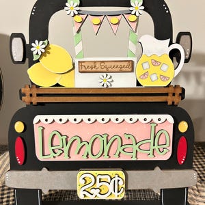 May include: A wooden lemonade stand shaped like a black truck with a pink sign that says "Lemonade" in green letters. The stand features lemons, a pitcher of lemonade, and a price tag of 25p.