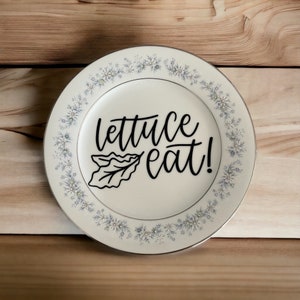 May include: A white ceramic dinner plate with a blue and white floral pattern around the edge. The plate has a black vinyl decal with the text "lettuce eat!" and a stylized lettuce leaf.
