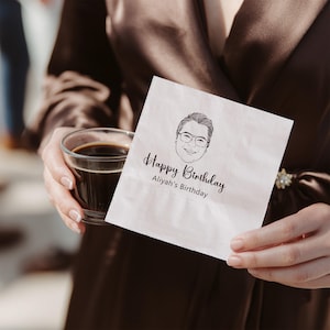Può includere: Un tovagliolo di carta bianco con un disegno a tratto nero di una persona con gli occhiali e la scritta "Happy Birthday Aliyah's Birthday" in corsivo nero. Il tovagliolo è tenuto da una persona con un abito marrone.