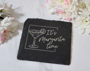 Può includere: Un sottobicchiere quadrato in ardesia nera con la scritta incisa "It's Margarita time". Il design include un bicchiere da margarita con ghiaccio e una fetta di lime. Fiori bianchi con accenti rosa sullo sfondo.