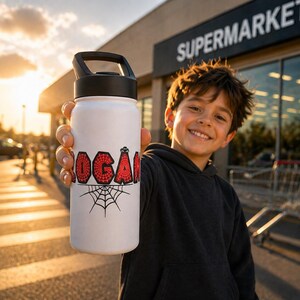 May include: White water bottle with a black handle and lid. The name "LOGAN" is printed in red, with a spiderweb design. A black spiderweb graphic is below the name. The bottle is held by a person in front of a supermarket.