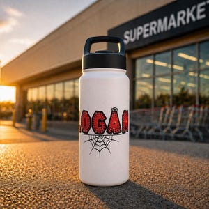 May include: White water bottle with a black handle and lid. The bottle features the name "LOGAN" in red, web-patterned lettering, with a spider and web graphic below. The bottle is standing outside a supermarket.