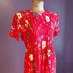 May include: A red floral print dress with short sleeves. The dress features a round neckline and pleats down the front. The floral pattern includes yellow, blue, and white flowers with green leaves. The dress is displayed on a mannequin.