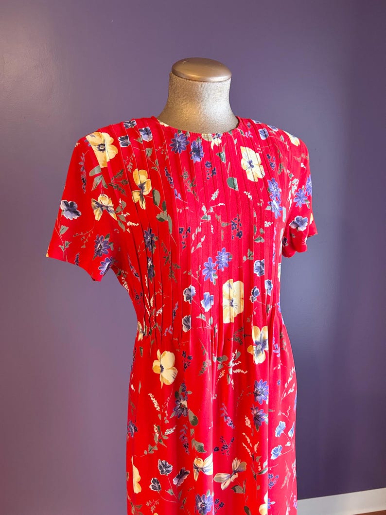 May include: A red floral print dress with short sleeves. The dress features a round neckline and pleats down the front. The floral pattern includes yellow, blue, and white flowers with green leaves. The dress is displayed on a mannequin.