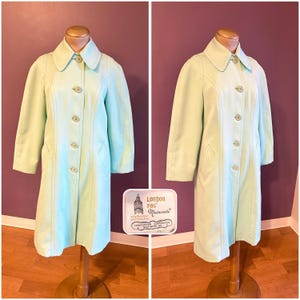 May include: A light green London Fog Maincoats trench coat. The coat has a classic design with a collar, button closure, and pockets. The coat is displayed on a wooden mannequin. The London Fog label is visible.