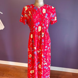 May include: A red floral print dress with short sleeves and a pleated bodice. The dress features a pattern of yellow and blue flowers. The dress is displayed on a mannequin. The background is a purple wall and a wooden floor.