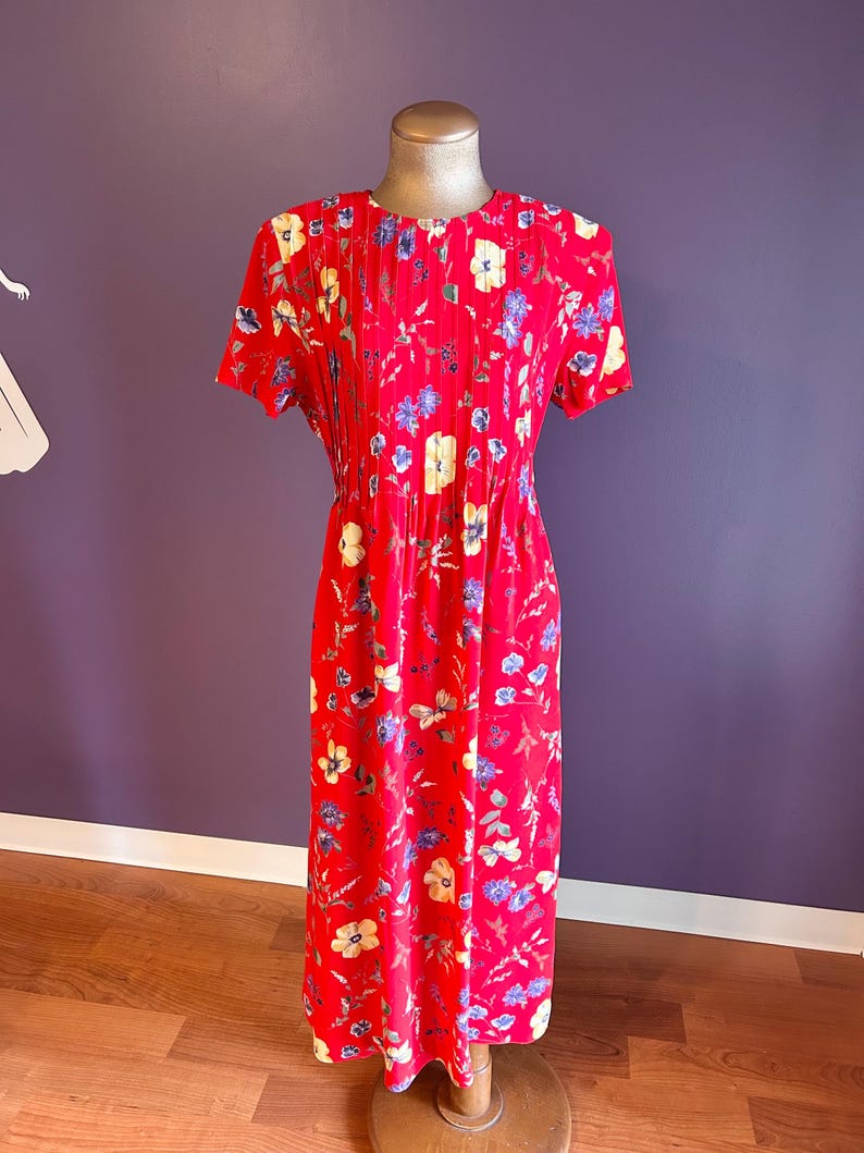May include: A red floral print dress with short sleeves and a pleated bodice. The dress features a pattern of yellow and blue flowers. The dress is displayed on a mannequin. The background is a purple wall and a wooden floor.