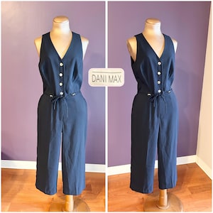 May include: Navy blue jumpsuit with a V-neckline and button-up front. The jumpsuit features a matching belt and wide-leg pants. The text "DANI MAX" is displayed on a white rectangular label. The outfit is displayed on a mannequin.