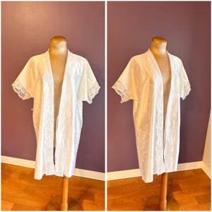 Może przedstawiać: Biały szlafrok z otwartym przodem i koronkowym wykończeniem na rękawach i krawędziach. Tkanina ma subtelny kwiatowy wzór. Szlafrok jest prezentowany na manekinie. Lekki, przewiewny element garderoby.