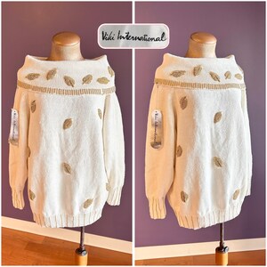Puede incluir: Un suéter de punto color blanco roto con un cuello ancho doblado y mangas largas. La prenda está decorada con motivos de hojas doradas con cuentas dispersas por la parte delantera y el cuello. Tiene una etiqueta de Viki International.