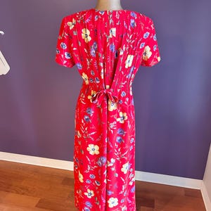 May include: A red floral print dress with short sleeves and a pleated back detail. The dress features a tie at the waist and is adorned with yellow, blue, and white flowers. The dress is displayed on a mannequin.