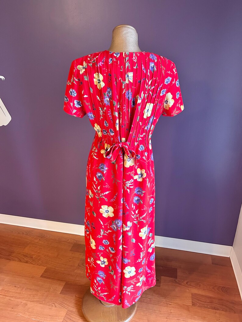 May include: A red floral print dress with short sleeves and a pleated back detail. The dress features a tie at the waist and is adorned with yellow, blue, and white flowers. The dress is displayed on a mannequin.