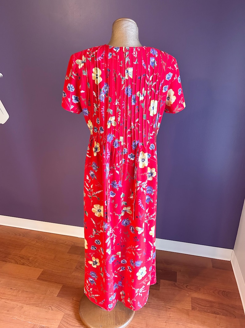May include: A red floral print dress with short sleeves and a pleated back. The dress features a pattern of yellow, blue, and white flowers. The dress is displayed on a mannequin.