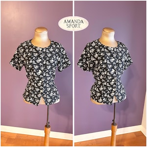May include: Black floral print blouse with short sleeves and a button-up front. The blouse is displayed on a mannequin. The text "AMANDA SPORT" is in a white oval in the upper center of the image.