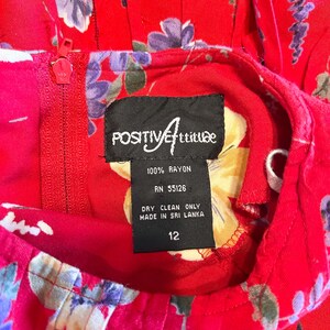 May include: A red floral dress with a zipper and a tag that reads "POSITIVE Attitude". The dress is made of 100% rayon and is a size 12. The tag also indicates that it was made in Sri Lanka and should be dry cleaned.