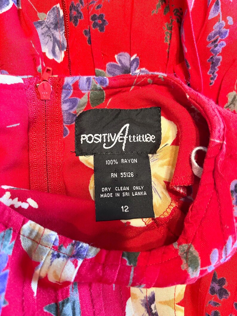 May include: A red floral dress with a zipper and a tag that reads "POSITIVE Attitude". The dress is made of 100% rayon and is a size 12. The tag also indicates that it was made in Sri Lanka and should be dry cleaned.