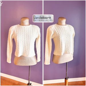 May include: Cream-coloured cable knit sweater from Liz Claiborne Petite Collection. The long-sleeved sweater is displayed on a mannequin against a purple background. The sweater has a crew neck and a cropped fit.