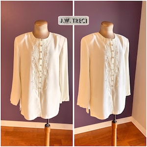 May include: Off-white long-sleeve blouse with a delicate lace panel down the front, secured by small, round buttons. The blouse has a classic, slightly loose fit and is displayed on a mannequin. The label reads "J.W. TRECI".