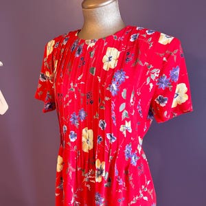May include: A red floral print dress with short sleeves. The dress features a round neckline with vertical pleats and is adorned with a pattern of yellow, blue, and white flowers and green leaves. The dress is displayed on a mannequin.