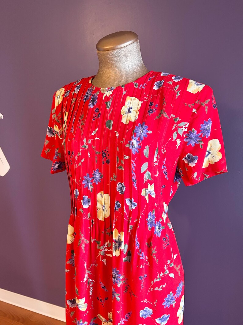 May include: A red floral print dress with short sleeves. The dress features a round neckline with vertical pleats and is adorned with a pattern of yellow, blue, and white flowers and green leaves. The dress is displayed on a mannequin.
