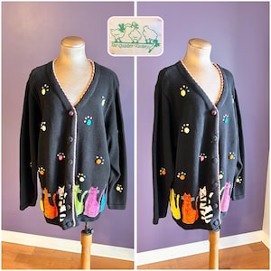 以下が含まれることがあります： Vネックの黒いカーディガン。カラフルな猫と足跡のデザインが特徴です。ピンクのトリムとボタン留めが付いています。左上にThe Quacker Factoryのロゴが見えます。