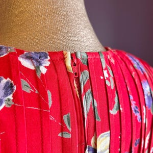 May include: Close-up of a red dress with a floral pattern. The dress features a zipper and vertical pleats. The floral design includes purple, white, and green accents. The dress is displayed on a mannequin.