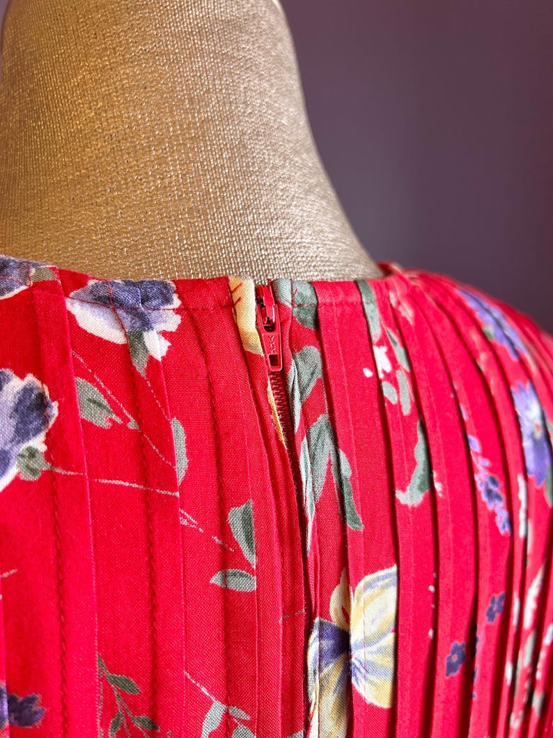 May include: Close-up of a red dress with a floral pattern. The dress features a zipper and vertical pleats. The floral design includes purple, white, and green accents. The dress is displayed on a mannequin.