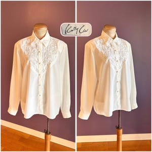 May include: A white long-sleeved button-down shirt featuring a decorative lace panel on the chest and a classic collar. The shirt includes a front button closure with small round buttons and a label displaying the text Cathy Che.