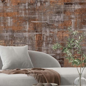 May include: A light gray sofa with a matching square throw pillow and a brown fringed throw blanket. A clear glass vase with green leafy branches and a small gray bowl sit in the foreground against a textured wall with rust and gray tones.
