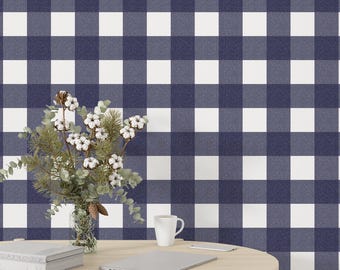 May include: A navy blue and white checkered pattern wallpaper. A wooden table holds a vase of flowers, a book, a laptop, a white mug, and a small white rectangle. The background is a large-scale gingham pattern.