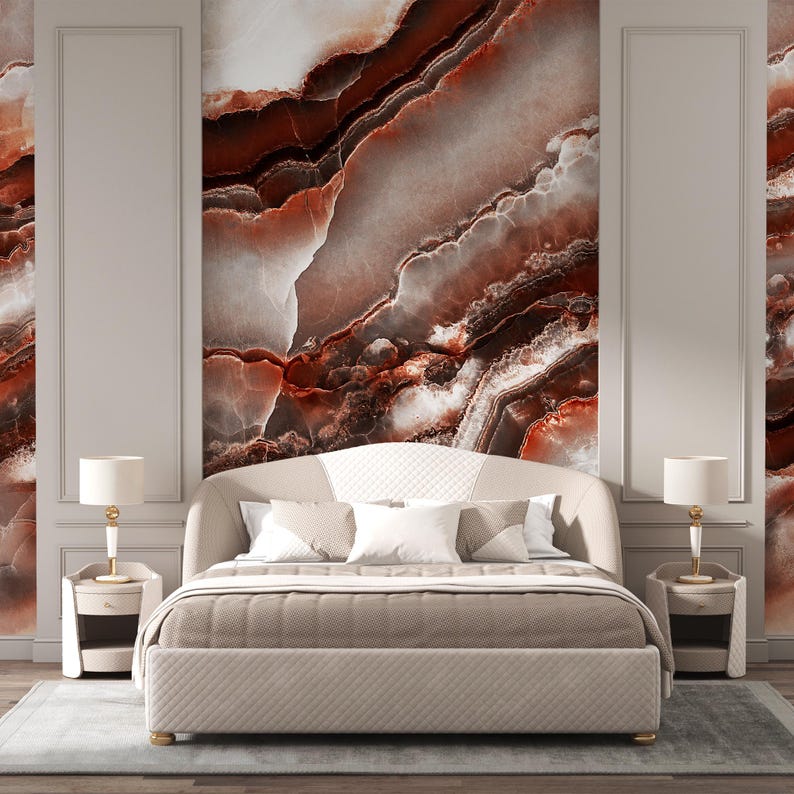 May include: A beige upholstered bed with a curved headboard featuring a quilted pattern, positioned between two matching beige nightstands. Each nightstand holds a white lamp with a gold base. The wall behind the bed displays a large marble mural.