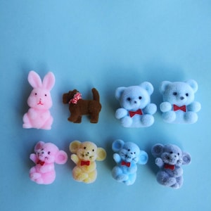 May include: A collection of small, fuzzy animal figurines on a light blue background. The figurines include a pink bunny, a brown dog with a red and white collar, and several teddy bears and mice in various colors, each with a small red bow tie.